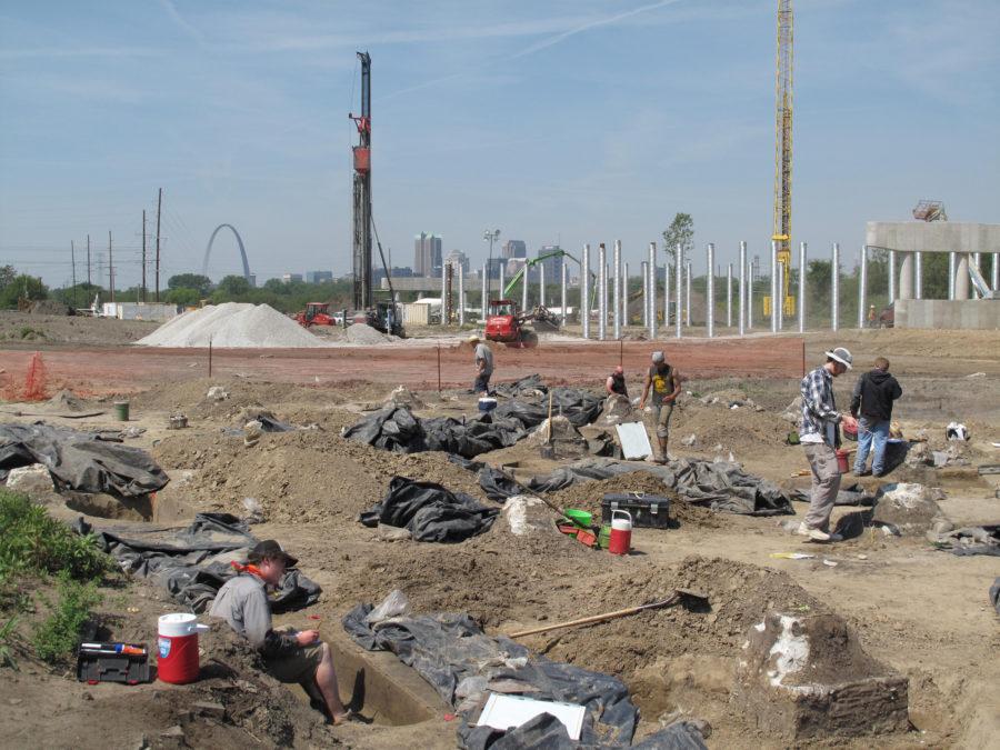 The University excavation crew works at the dig site while the St. Louis skyline looms in background.
