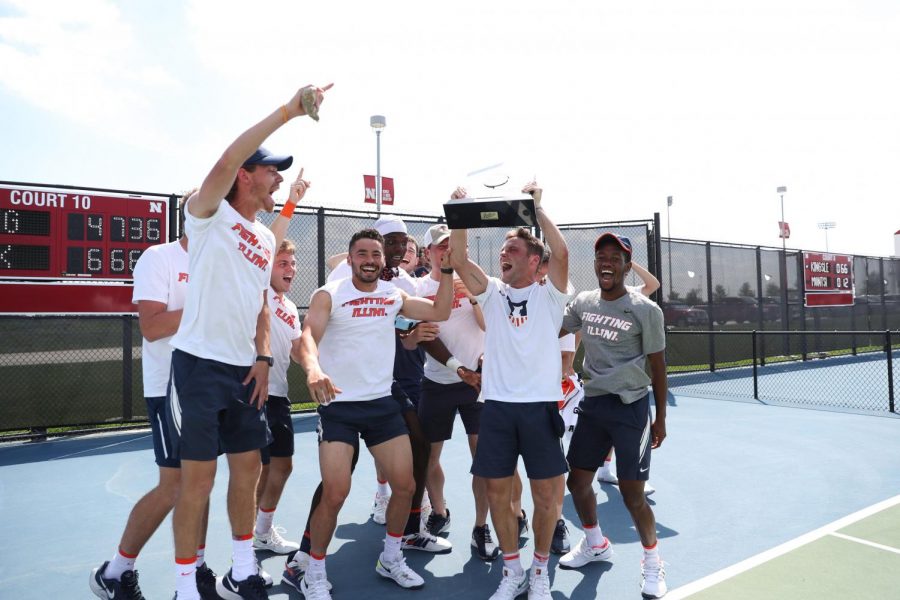 The men’s tennis team celebrates on a court with their Big Ten Championship trophy.
