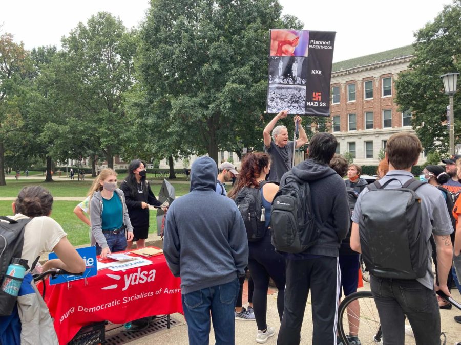 A man stands next to a democratic table holding a sign sharing that Planned Parenthood is killing humans like the Nazis or the KKK. Students debated against him saying that he should not compare the two. 
