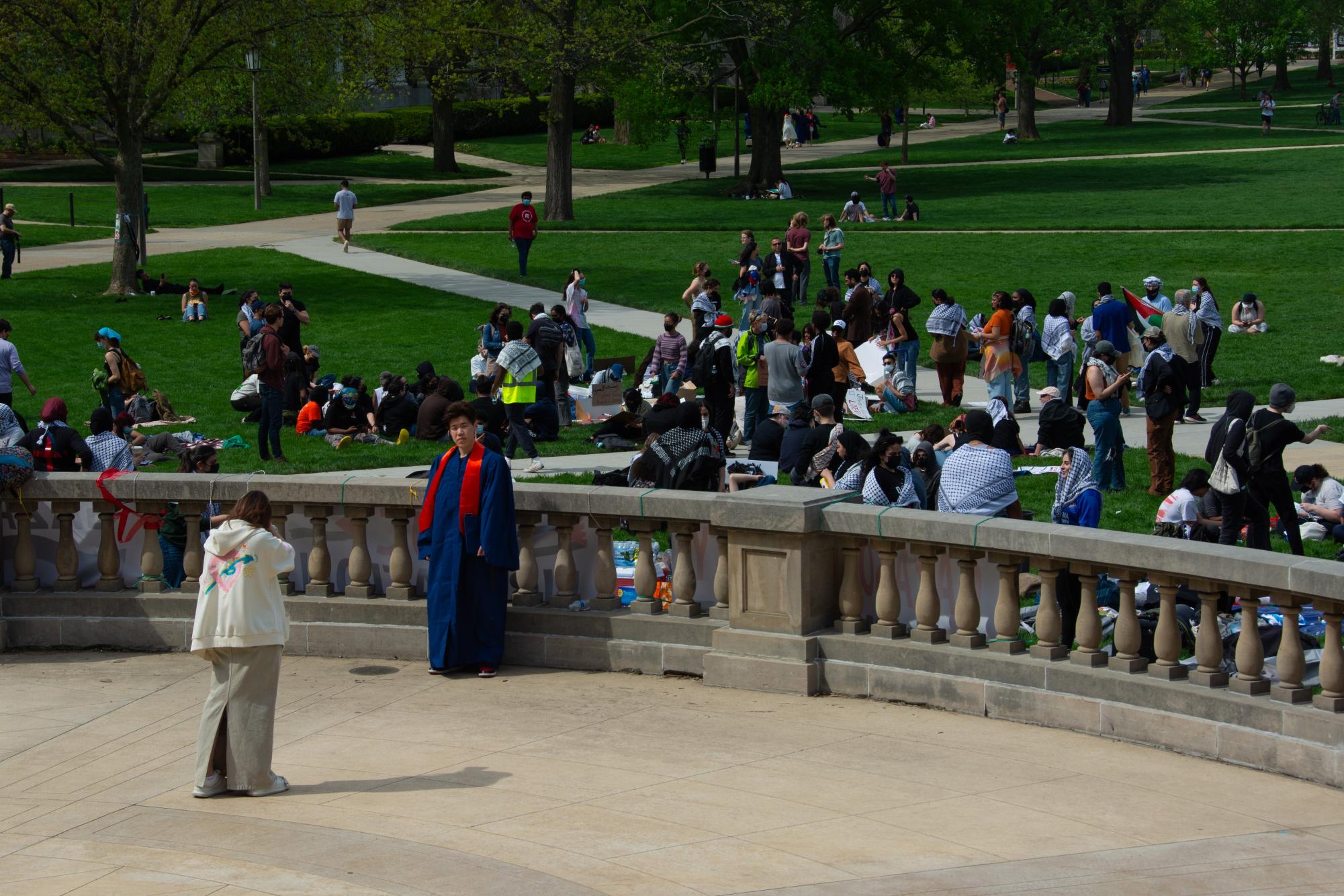 Students take graduation photos as the protesters assemble on the Main Quad in the background.