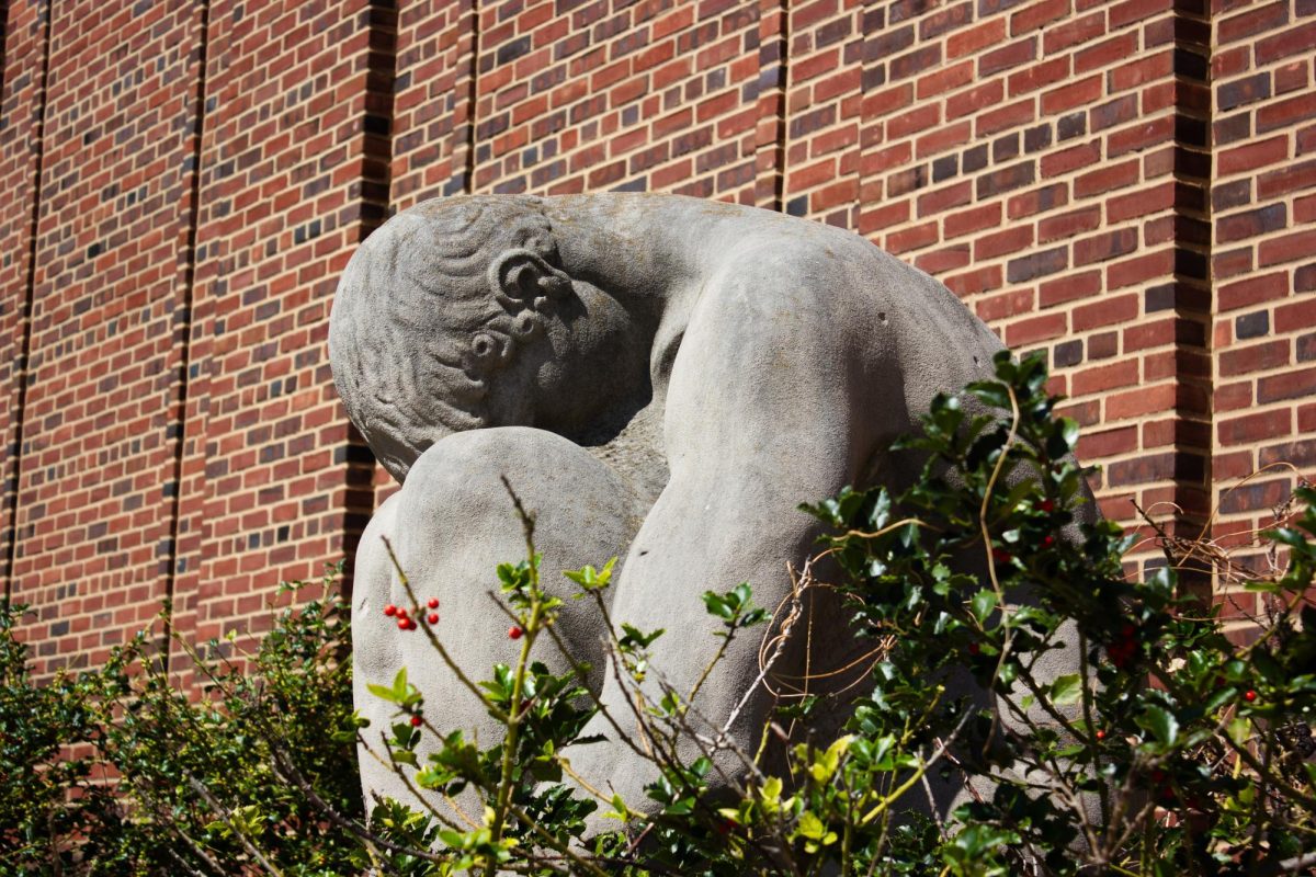 A stone statue depicting a hunched person situated behind Foellinger Auditorium and surrounded by greenery.
