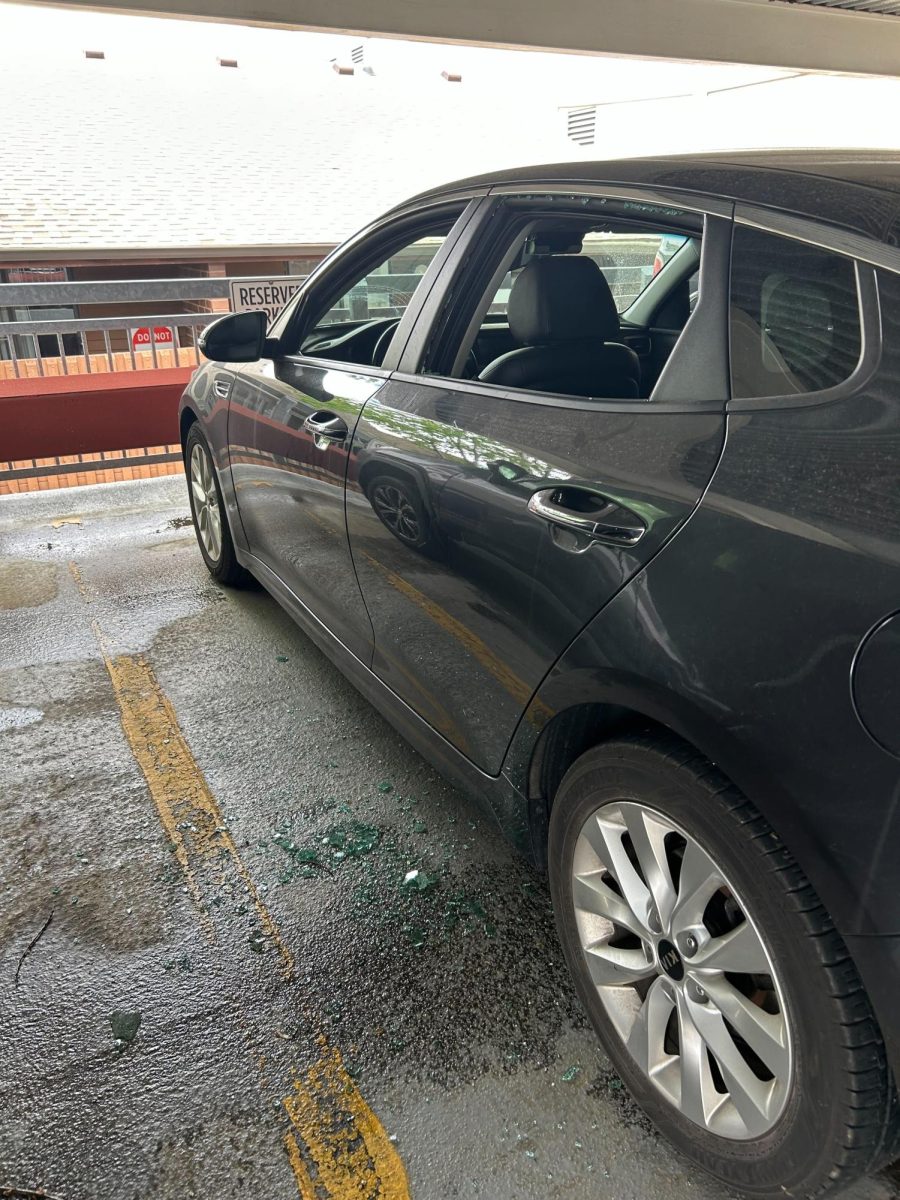 A car parked in a parking lot with its window shattered and glass on the ground. 