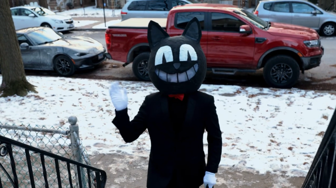 A figure wearing a cat head stands on a front porch in the music video for Jenny Scara's latest release, “Black Cat.”